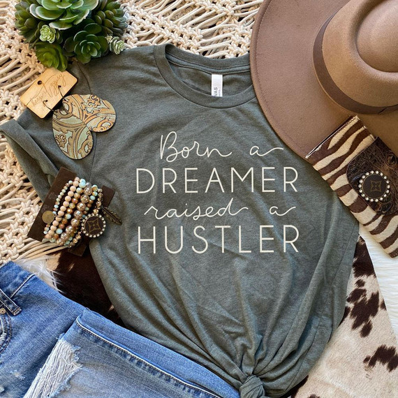 Born a Dreamer Screen Print Heat Transfer, gray T-shirt with the text 'Born a DREAMER raised a HUSTLER' printed on it, surrounded by jewelry, a hat, and decorative items.