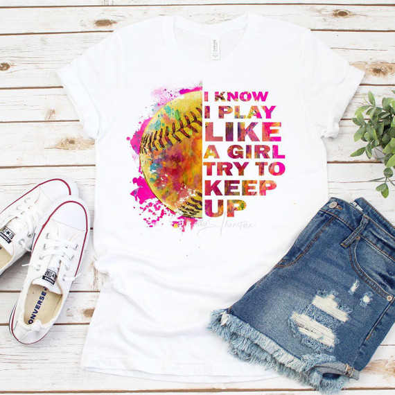 I Know I Play Like a Girl Try To Keep Up sublimation transfer t-shirt with colorful baseball graphic design, laid flat on a white wooden surface with sneakers and denim shorts nearby.
