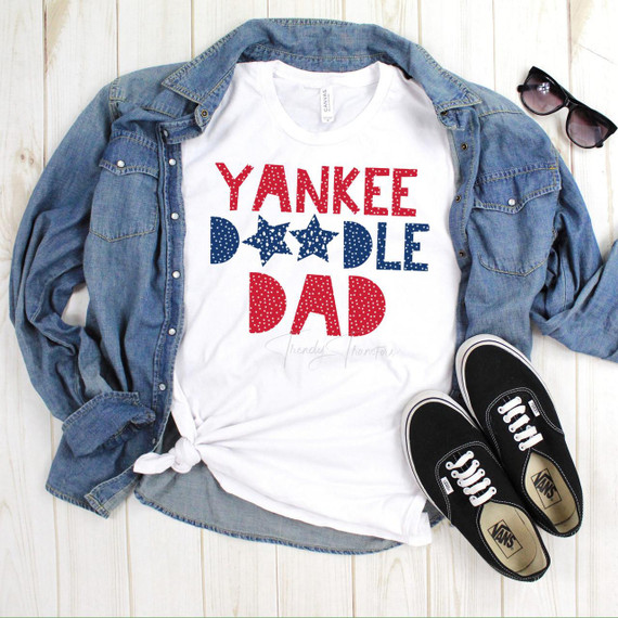 Yankee Doodle Dad family set sublimation transfer, a white T-shirt with red and blue patriotic star-themed text design laid out beside a denim shirt, black Vans shoes, and sunglasses on a wooden background.