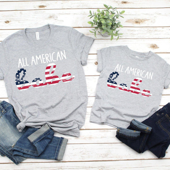 All American Babe YOUTH & INFANT - FINAL STOCK Screen Print Heat Transfer T-shirts for children featuring patriotic American themed design with red, white, and blue stars and stripes, displayed on a white wooden background with denim jeans and a green plant.