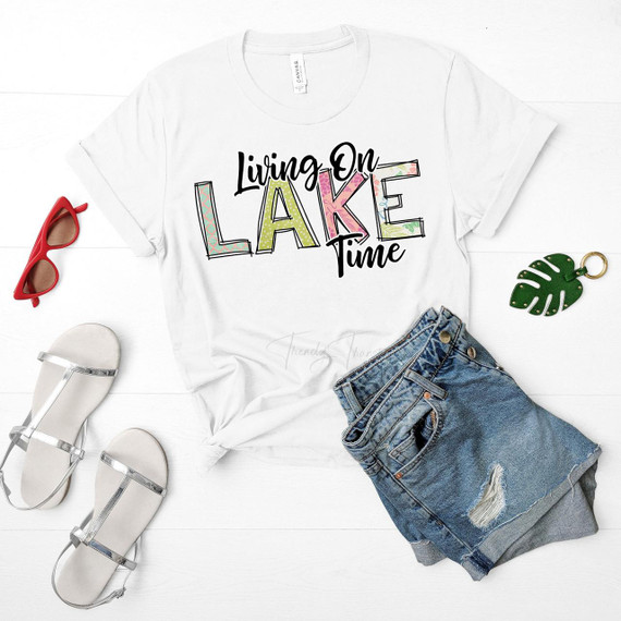 Living On Lake Time Sublimation Transfer T-shirt mockup with colorful, decorative text, paired with casual denim shorts, white sandals, red sunglasses, and a green fridge magnet leaf, laid out on a white wooden surface.
