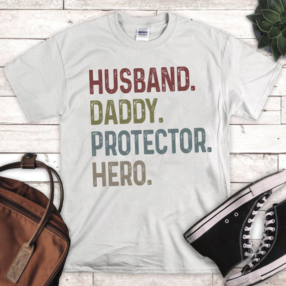 Husband Daddy Protector Hero Sublimation Transfer, T-shirt with colorful stamped text design displayed on wooden background with backpack, sneakers, and plant accessories.