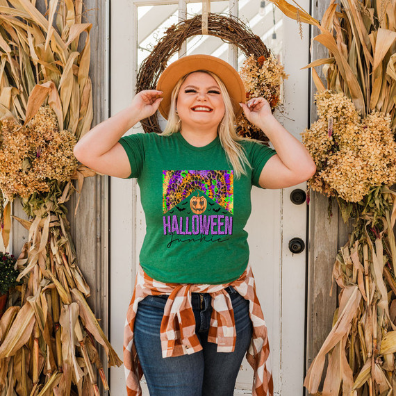 Halloween Junkie DTF Heat Transfer, vibrant and colorful design featuring a pumpkin and the word Halloween on a green t-shirt worn by a smiling woman in fall attire, surrounded by autumn decorations.