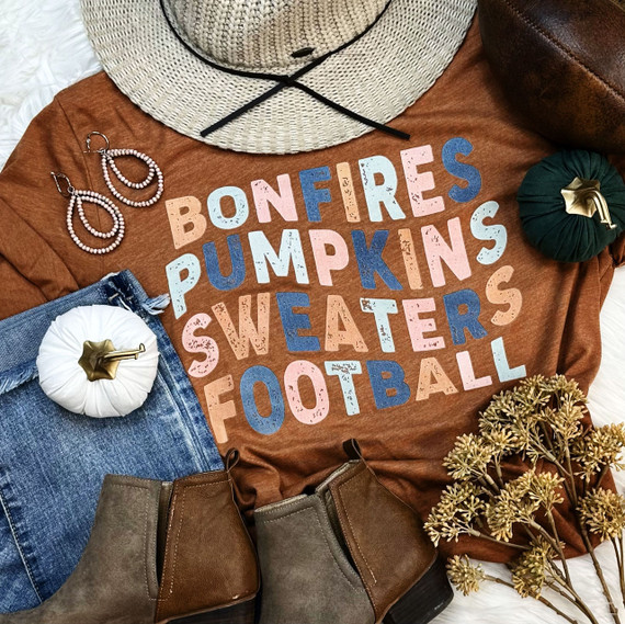 Bonfires and Football Screen Print Heat Transfer, colorful printed text on a brownish-orange t-shirt surrounded by autumn-themed items like hats, jewelry, pumpkins, boots, and dried flowers.