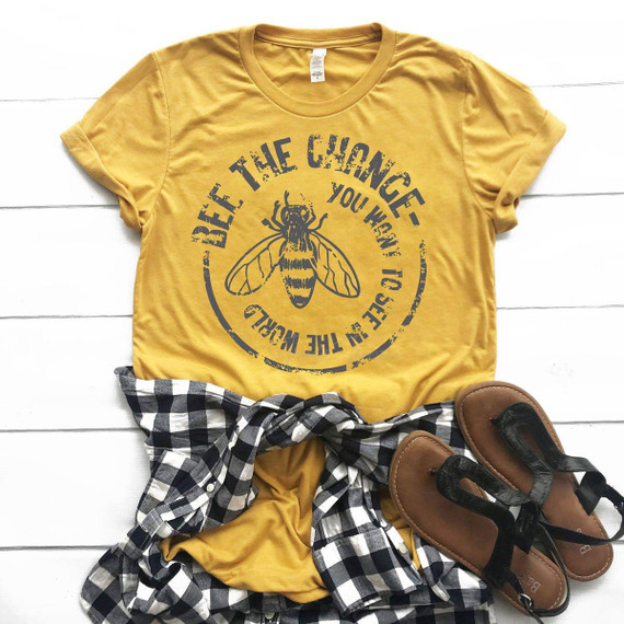 Bee The Change Screen Print Heat Transfer, yellow t-shirt with a bee graphic and motivational text, black and white checkered shirt, and brown sandals.