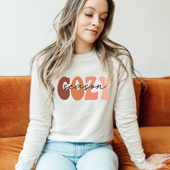Cozy Season Screen Print Heat Transfer, woman wearing a sweatshirt with 'cozy season' graphic design sitting on an orange sofa.