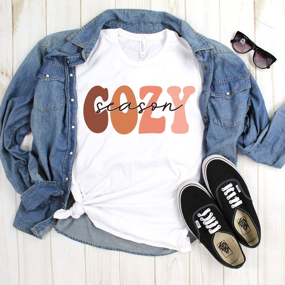 Cozy Season Sublimation Transfer, white T-shirt with 'COZY season' text in brown and pink, paired with denim shirt, black Vans shoes, and sunglasses on a light wooden background.