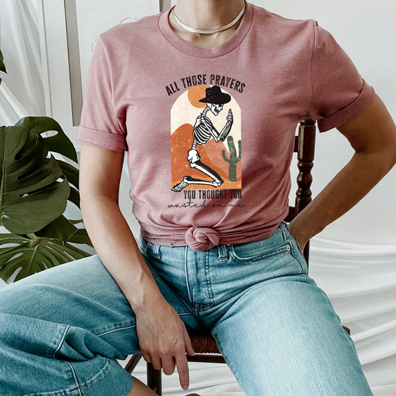 All Those Prayers Screen Print Heat Transfer T-Shirt featuring a skeleton praying with cactus and sunset background.
