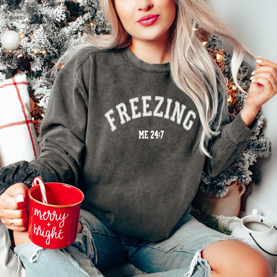 Freezing 24:7 Screen Print Heat Transfer, a woman wearing a dark gray sweatshirt with the text 'FREEZING ME 24:7', holding a red mug with 'merry & bright' written on it, sitting near a Christmas tree with ornaments and decorations.