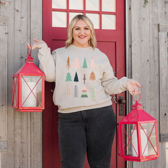 Boho Trees Screen Print Heat Transfer, a woman holding two red lanterns and wearing a sweatshirt with colorful boho-style tree prints, standing in front of a rustic wooden wall and red door.