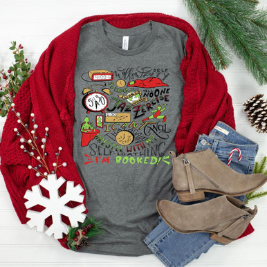 I'm Booked DTF Transfer, Christmas-themed gray T-shirt with colorful travel and holiday icons, displayed with red sweater, jeans, and boots on a festive background.