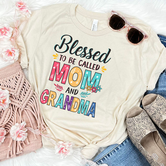 Blessed To Be Called Mom And Grandma DTF Transfer, colorful and decorative t-shirt with floral and heart accents, displayed alongside pink flowers, sunglasses, shoes, and blue jeans for a stylish mom and grandma gift.
