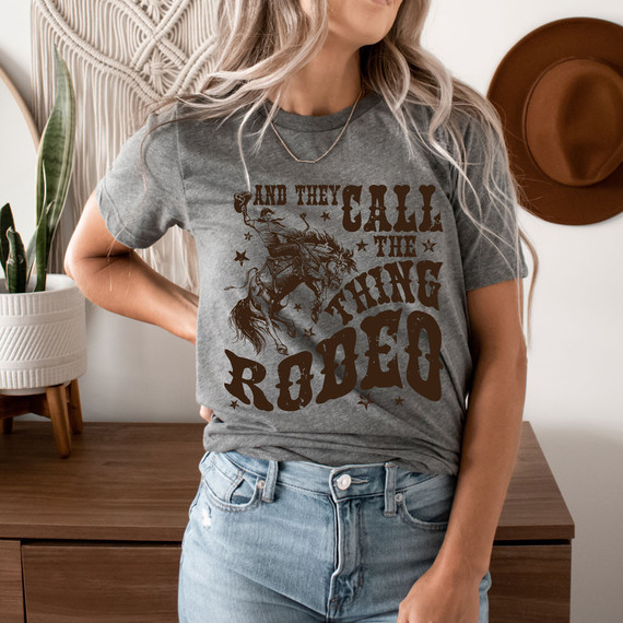 And They Call The Thing Rodeo Screen Print Heat Transfer, a humorous rodeo-themed graphic on a gray t-shirt worn by a woman.