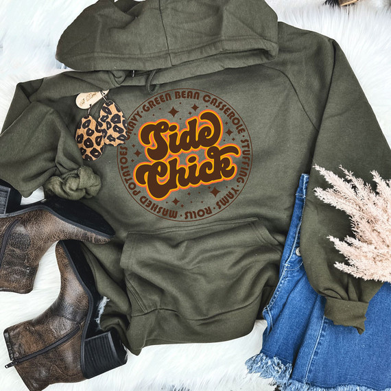 Side Chick DTF Transfer, a colorful circular design with the text 'Side Chick' in bold, yellow and orange fonts, placed on a dark green hoodie surrounded by casual fashion accessories.