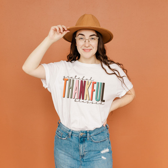 Grateful Thankful Blessed DTF Transfer, woman wearing a white t-shirt with colorful text design and a tan hat against an orange background.