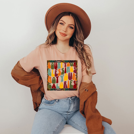 Count Your Blessings Not Your Calories DTF Transfer, a colorful graphic design on a woman's pink t-shirt with a woman wearing a brown hat and jacket.