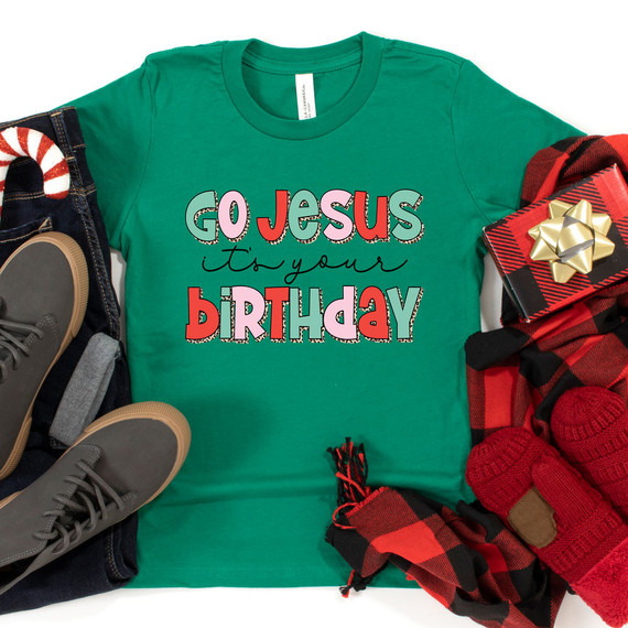 Go Jesus It's Your Birthday DTF Transfer, colorful t-shirt with holiday-themed text and designs, surrounded by wrapped gift box, plaid scarf, red knit gloves, shoes, and Christmas decorations.