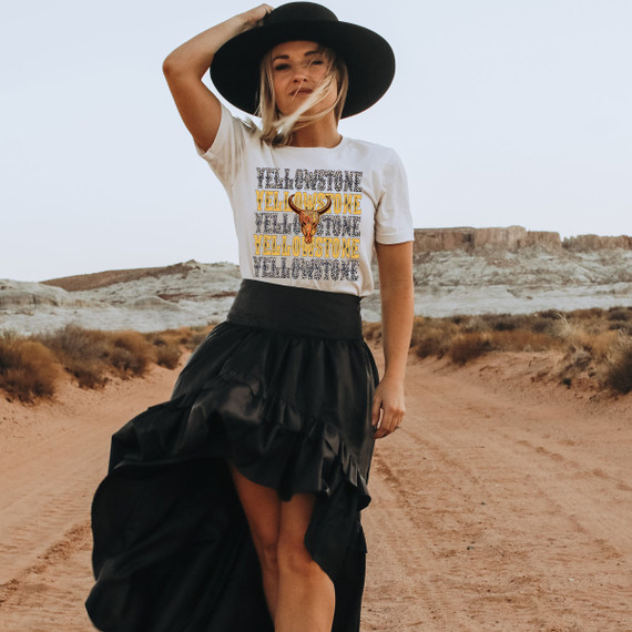 Yellowstone Stacked Screen Print Heat Transfer, a woman wearing a white t-shirt with a layered Yellowstone design and a deer skull graphic, standing in a desert landscape with rocky formations and sparse vegetation.