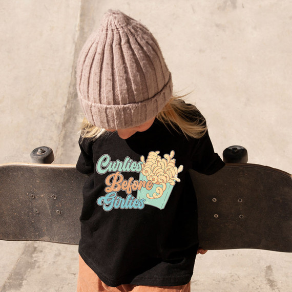 Curlies Before Girlies DTF Transfer, a colorful graphic design of a carton of curly fries with the text "Curlies Before Girlies" on a black t-shirt worn by a child holding a skateboard.