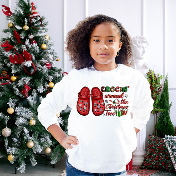 Crocin' Around The Christmas Tree DTF Heat Transfer, a girl wearing a white sweatshirt with a Christmas-themed design featuring red shoes, candy cane, gifts, and festive text, standing in front of a decorated Christmas tree.