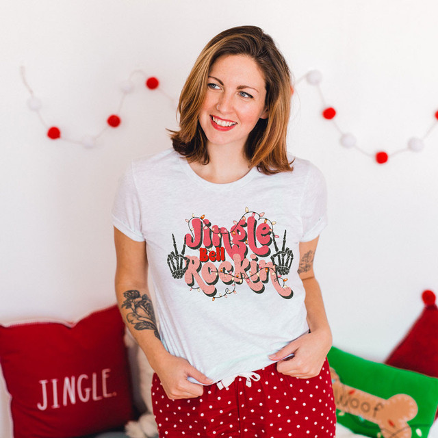 Jingle Bell Rockin' #2 Screen Print Heat Transfer, a woman showcasing a Christmas-themed t-shirt with the words "Jingle Bell Rockin'" decorated with holiday lights and skeletal hands, in a festive room.