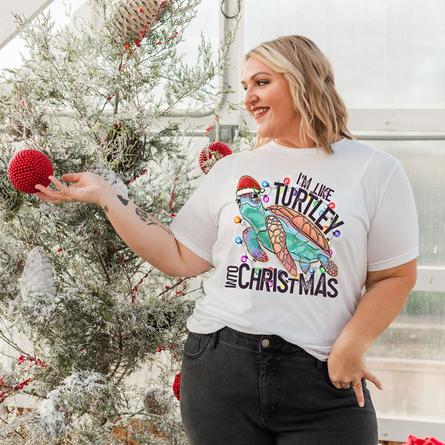 I'm Like Turtley Into Christmas DTF Heat Transfer, woman wearing a Christmas-themed t-shirt featuring a colorful turtle wearing a Santa hat, holiday decorations, Christmas tree, and festive ornaments.