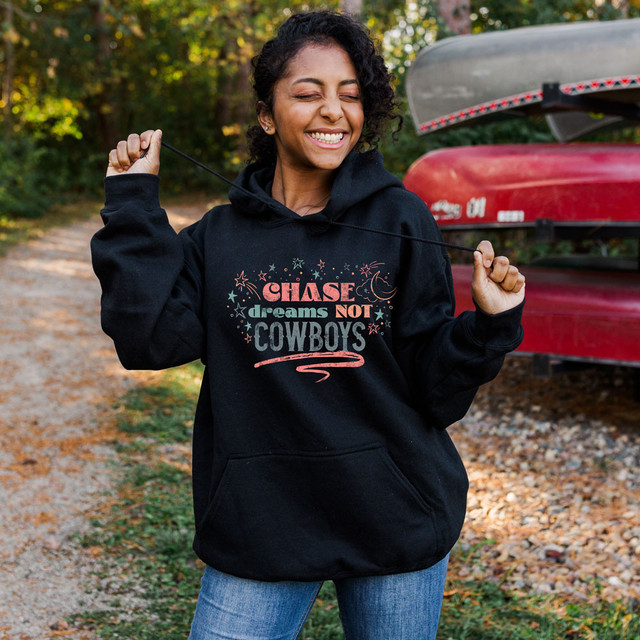 Chase Dreams Not Cowboys Screen Print Heat Transfer, a woman smiling outdoors wearing a black hoodie with colorful text and graphic design.
