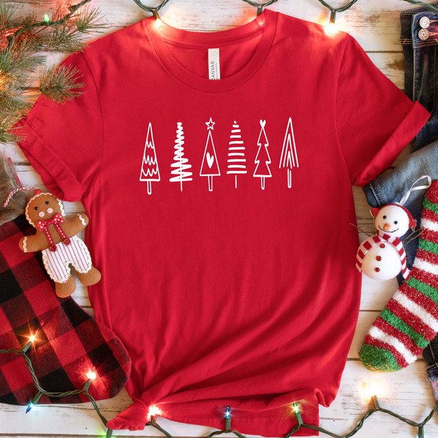 White Christmas Trees Screen Print Heat Transfer, a festive red t-shirt with a white Christmas trees design, surrounded by Christmas decorations including plush holiday characters and string lights.