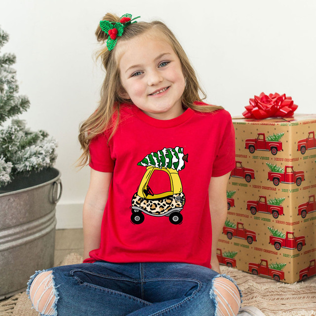 Leopard Tree Car DTF Heat Transfer, a colorful and playful heat transfer design on a red t-shirt worn by a smiling young girl, with Christmas decorations including a decorated Christmas tree and a wrapped gift box in the background.
