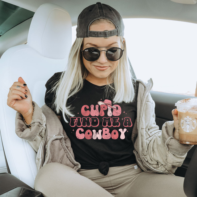 Cupid Find Me A Cowboy DTF Heat Transfer, woman wearing a black T-shirt with pink lettering and graphic, sitting in car with Starbucks drink.