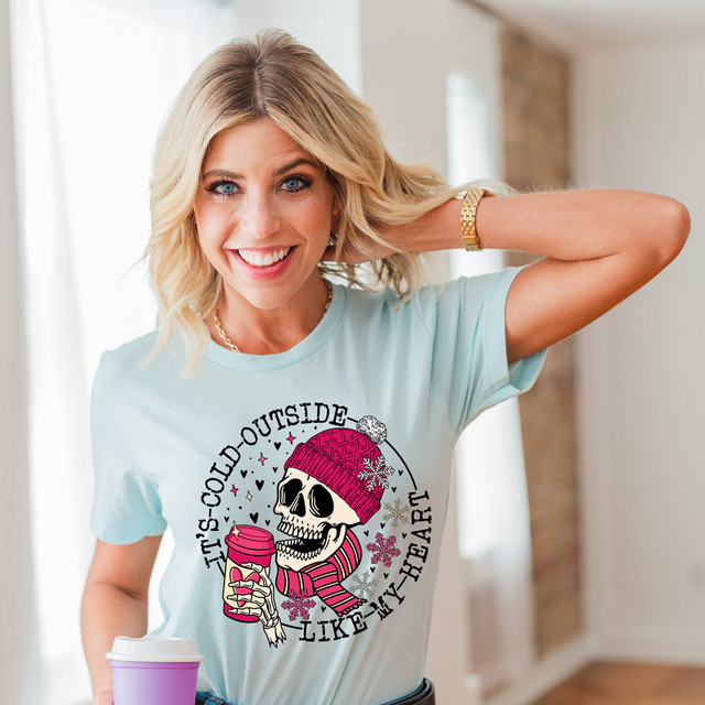 It's Cold Outside Like My Heart DTF Heat Transfer, a woman wearing a light blue t-shirt with a graphic of a skull wearing a pink hat and scarf holding a coffee cup.