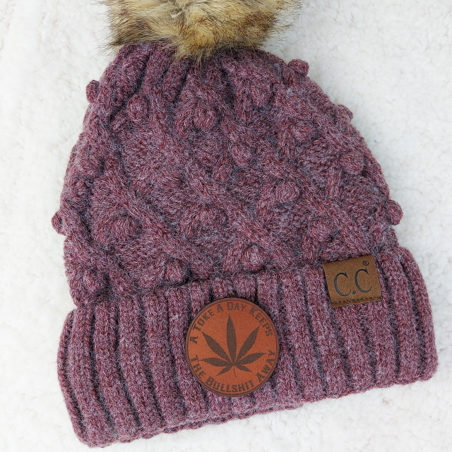 Product A Toke A Day Keeps The Bullshit Away Leather Patch, a knitted winter hat with a brown leather patch featuring the text and a cannabis leaf logo, and a small black leather logo tag with the initials C.C.