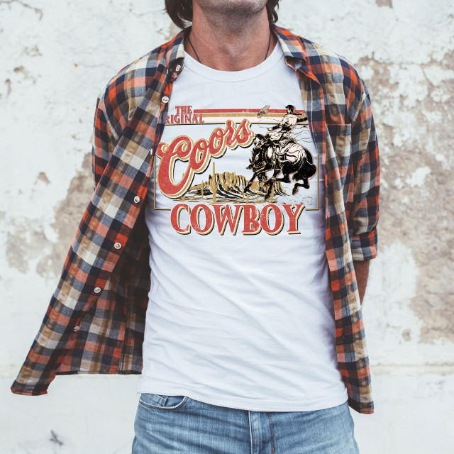 The Original Cowboy Screen Print Heat Transfer, graphic of a cowboy riding a horse with desert background on a white T-shirt worn by a man.