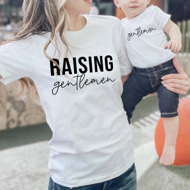 Gentleman DTF Heat Transfer, white T-shirt with 'RAISING gentleman' printed on it being worn by a woman holding a child in an outdoor setting.