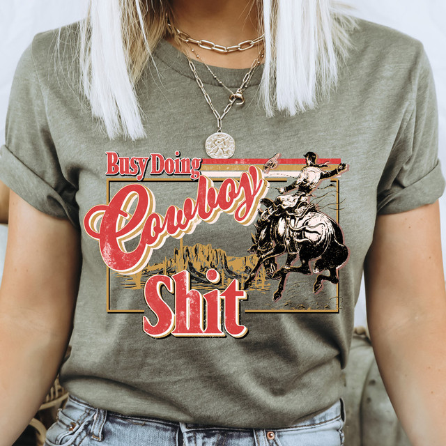 Busy Doing Cowboy Shit DTF Heat Transfer, humorous iron-on graphic design featuring a cowboy riding a bucking horse with a desert landscape background.