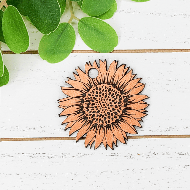 Sunflower Leather Keychain, a handcrafted orange sunflower-shaped leather keychain with black accents and a small circular hole for attaching keys, placed on a white wooden surface with green leaves nearby.