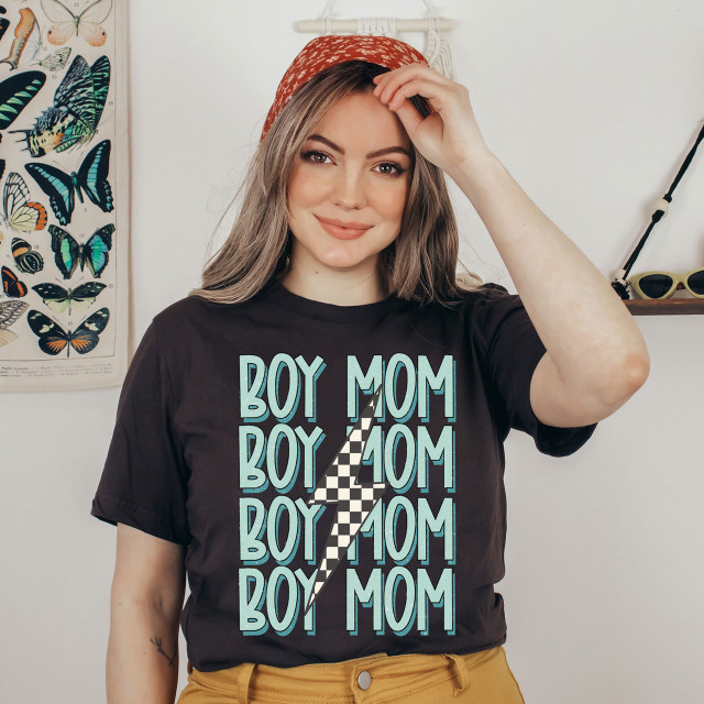 Boy Lightening Bolt Mom DTF Heat Transfer, woman wearing a black T-shirt with a multiple blue 'Boy Mom' text and lightning bolt design, indoor setting with butterfly poster and sunglasses in background.