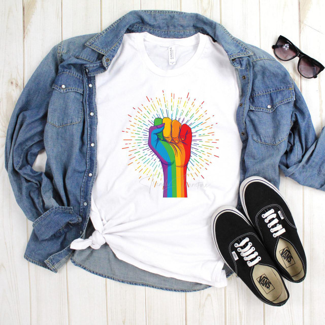 Pride Fist Rainbow Burst Sublimation Transfer, colorful rainbow fist design on a white T-shirt, laid on a wooden surface with denim jacket, black sneakers, and sunglasses.