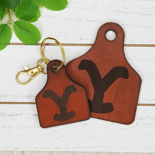 Yellowstone Leather Cattle Tag Keychain, leather keychain designed to resemble a cattle tag with a dark brown 'Y' logo, attached to a gold keyring and clip on a white wooden background with green leaves.