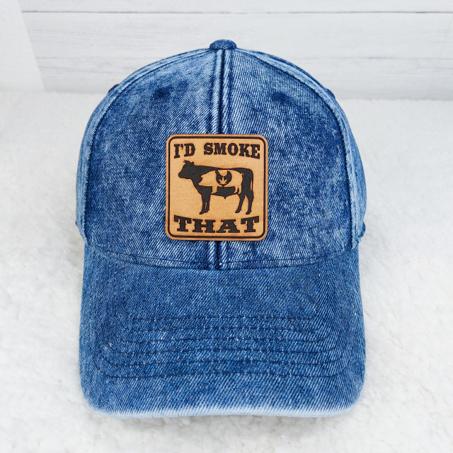I'D SMOKE THAT Leather Patch, a patch featuring a pot leaf design on a cow silhouette with the text I'D SMOKE THAT, sewn onto a blue denim baseball cap.