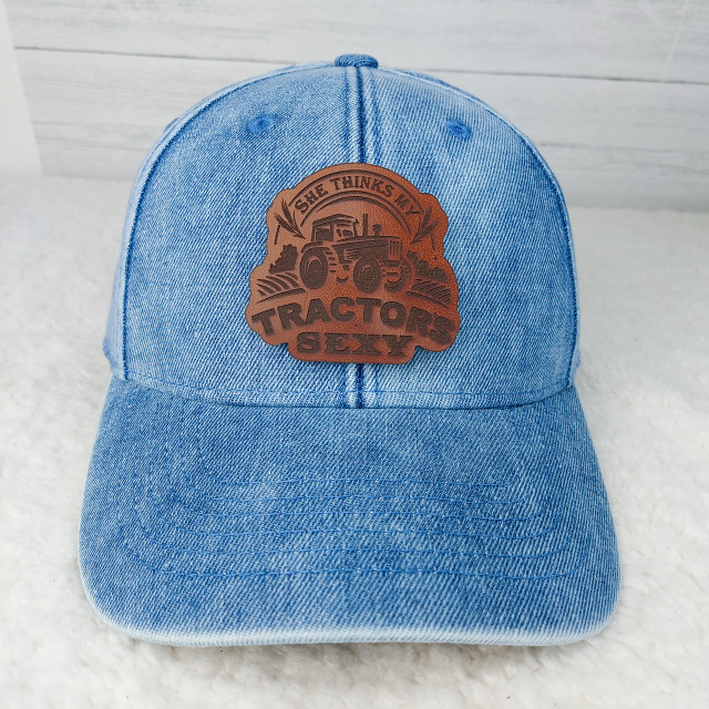 She Thinks My Tractor's Sexy Leather Patch baseball cap, featuring a brown leather patch with tractor graphic and text, on a light blue denim cap.