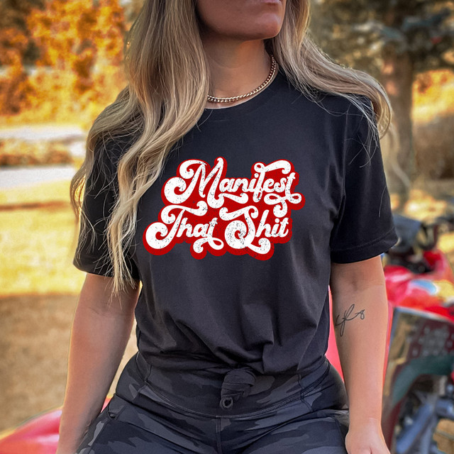 Manifest That Shit Distressed DTF Heat Transfer, a bold graphic design with white textured text outlined in red on a black t-shirt.
