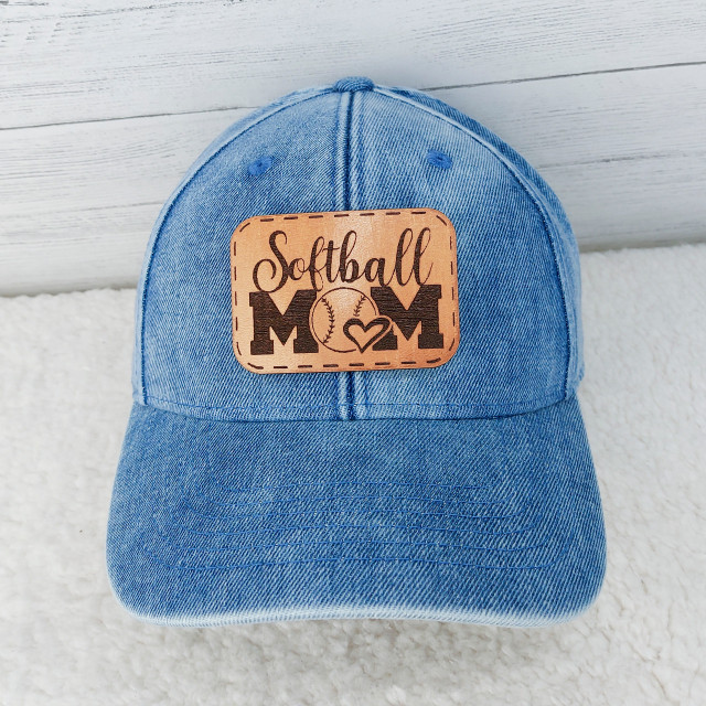 Softball Mom Leather Hat Patch, blue denim baseball cap with a tan leather patch embroidered with the words 'Softball Mom' and a baseball graphic surrounded by a stitched border.