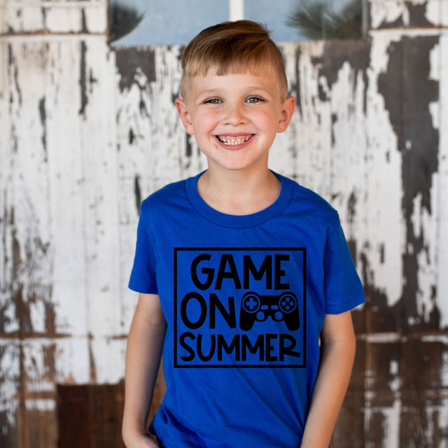Game On Summer DTF Heat Transfer, close-up of a smiling young boy wearing a bright blue t-shirt with a black game controller design and text, standing in front of a rustic wooden background.