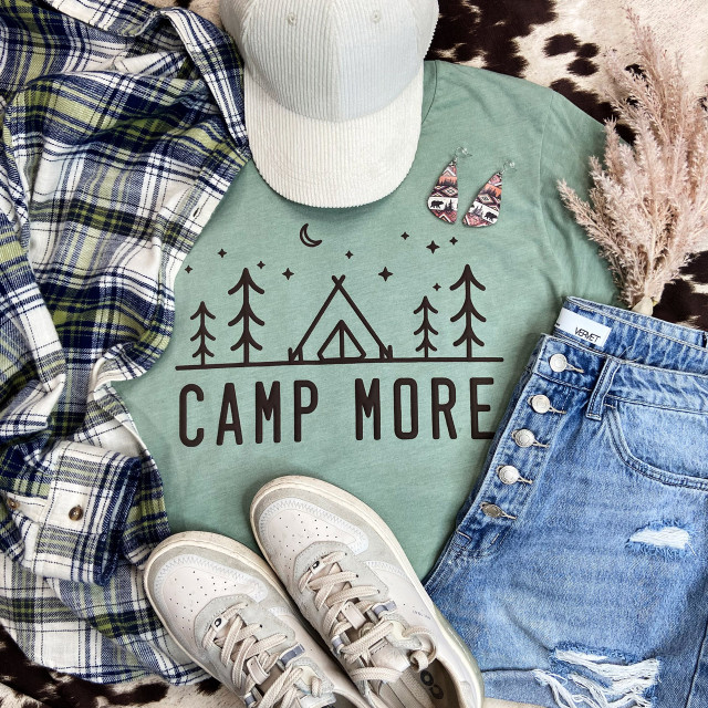 Camp More PUFF Screen Print Heat Transfer, image of a green t-shirt with camping-themed graphic, paired with white sneakers, denim shorts, a plaid shirt, and accessories including earrings and a hat, displayed with pink pampas grass.