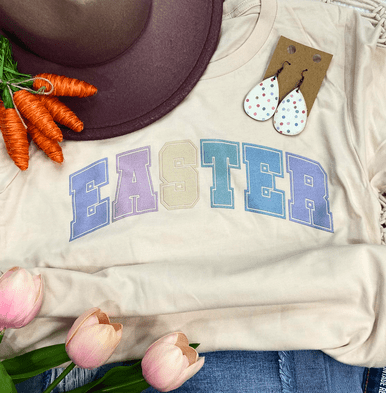 Easter Varsity Letters Screen Print Heat Transfer, a close-up view of pastel-colored varsity-style letters spelling 'EASTER' on a cream-colored t-shirt, decorated with jewelry and flowers.