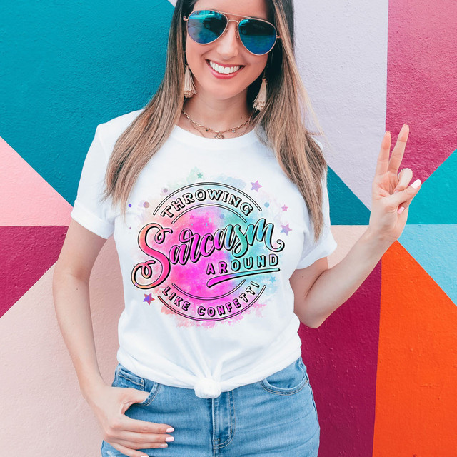 Throwing Sarcasm Around Like Confetti DTF Heat Transfer, woman wearing white T-shirt with colorful sarcastic quote design and rainbow background, standing in front of a vibrant mural wall.