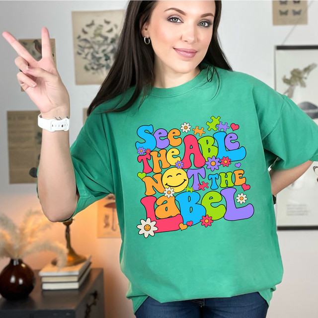See The Able Not The Label DTF Heat Transfer, colorful graphic T-shirt featuring a retro-style psychedelic design with flowers, hearts, and a smiling face.