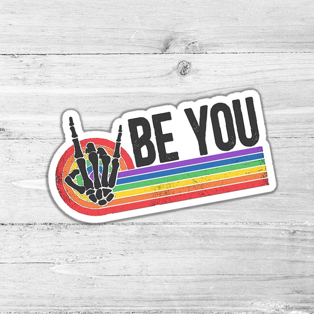 Be You Die Cut Sticker, colorful skeleton hand making a peace sign with rainbow stripes and the text 'BE YOU' on a white wooden background.