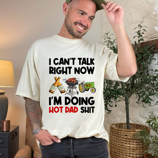 I Can't Talk Right Now I'm Doing Hot Dad Shit TDT Heat Transfer, Man wearing a T-shirt with BBQ grill, beer bottles, and tractor graphic, smiling in home setting.